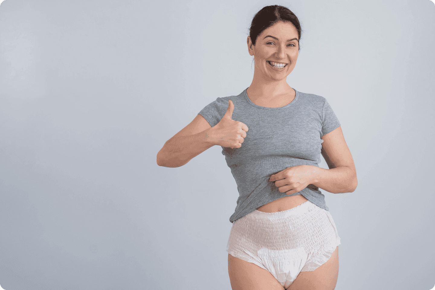 A smiling woman in a grey t-shirt and adult nappy gives a thumbs-up gesture. She holds up a corner of her shirt slightly to reveal the waistband of th