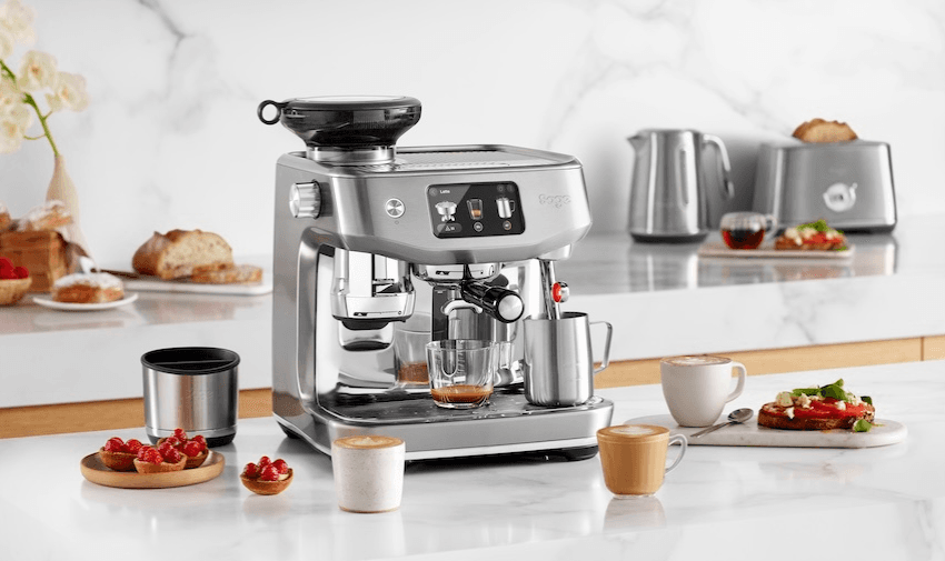 Modern silver Sage espresso machine on a marble kitchen counter with fresh bread, fruit, and cappuccinos, alongside matching kettle and toaster. Premium coffee machines for home brewing – mobility and kitchen appliance discounts for disabled people.