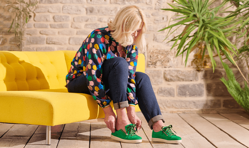 A woman sitting on a bright yellow sofa tying the laces of vibrant green trainers. She is wearing a colourful polka dot blouse and dark jeans. The setting features a rustic stone wall and indoor plants. Shopping Discounts for Disabled People.
