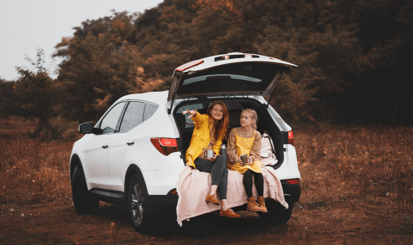 Two young girls in matching yellow coats sitting in the boot of a white SUV parked in nature, enjoying a cosy outdoor moment with blankets and hot drinks, representing family travel comfort and vehicle leisure use.