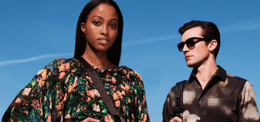 A woman with long straight hair wearing a floral print dress and a crossbody bag stands in the foreground. A man with short dark hair wearing sunglasses and a patterned shirt with a backpack stands slightly behind her. The sky is clear and blue.