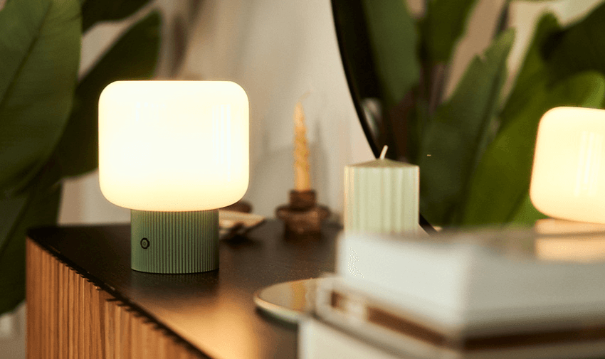 Softly glowing bedside lamp on a black wooden table beside a mirror, styled with decorative candles and leafy green plants, highlighting affordable home lighting options and energy-efficient solutions for disabled households in the UK.