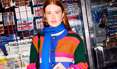 Young woman with striking red hair wearing a colourful striped jumper and vibrant blue scarf, standing outside a London newsagent showcasing modern, sustainable fashion by Omnes