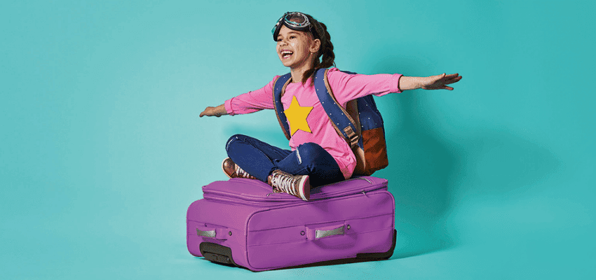 A young girl, full of joy, wearing a pink star shirt and sitting playfully on a purple suitcase, symbolising family travel and fun. 