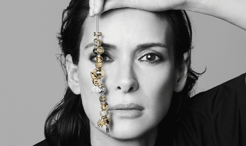 Woman holding a gold and silver Pandora charm bracelet vertically across her face, showcasing detailed jewellery design. High-contrast black and white portrait with a luxury fashion tone. 