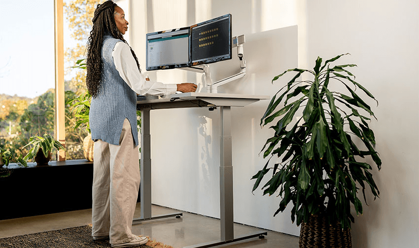 Black woman using a standing desk with dual monitors in a bright home office filled with greenery, demonstrating mobility-friendly work setups and accessible office furniture discounts for disabled people across the UK.
