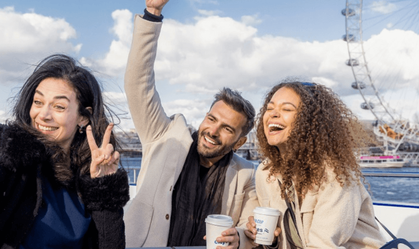 Three people enjoying a boat ride on a sunny day, holding coffee cups and smiling. London Eye is visible in the background against a partly cloudy sky.