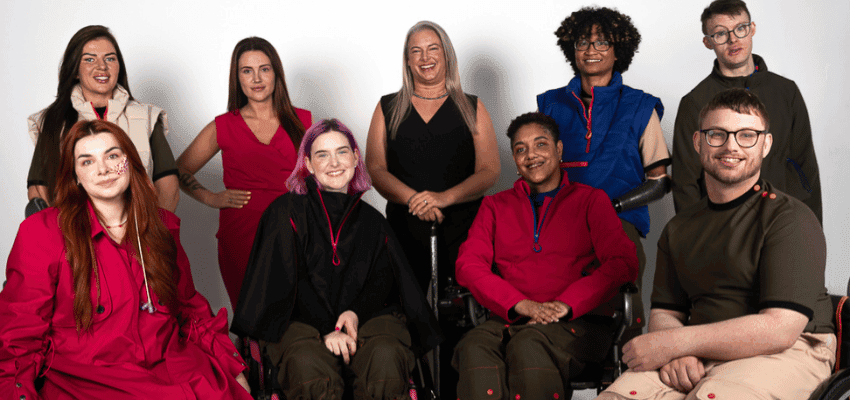 A diverse group of nine individuals, some in wheelchairs, are smiling and posing together. They are dressed in colourful and stylish clothing against a plain backdrop, conveying a sense of unity and inclusion.