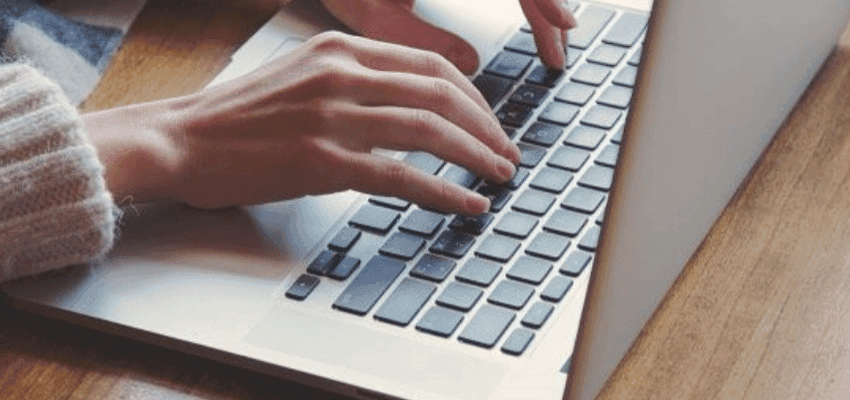 Close-up of a person's hands typing on a silver laptop with black keys. The person is wearing a light-coloured, knitted sweater and is seated at a wooden surface. The lighting is soft, creating a comfortable and relaxed atmosphere.