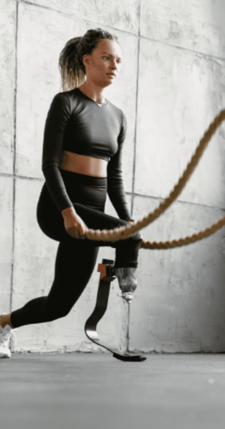 A woman with a prosthetic leg performing battle rope exercises in a minimalist gym, highlighting inclusivity and fitness. Discover disabled fitness discounts with Purpl