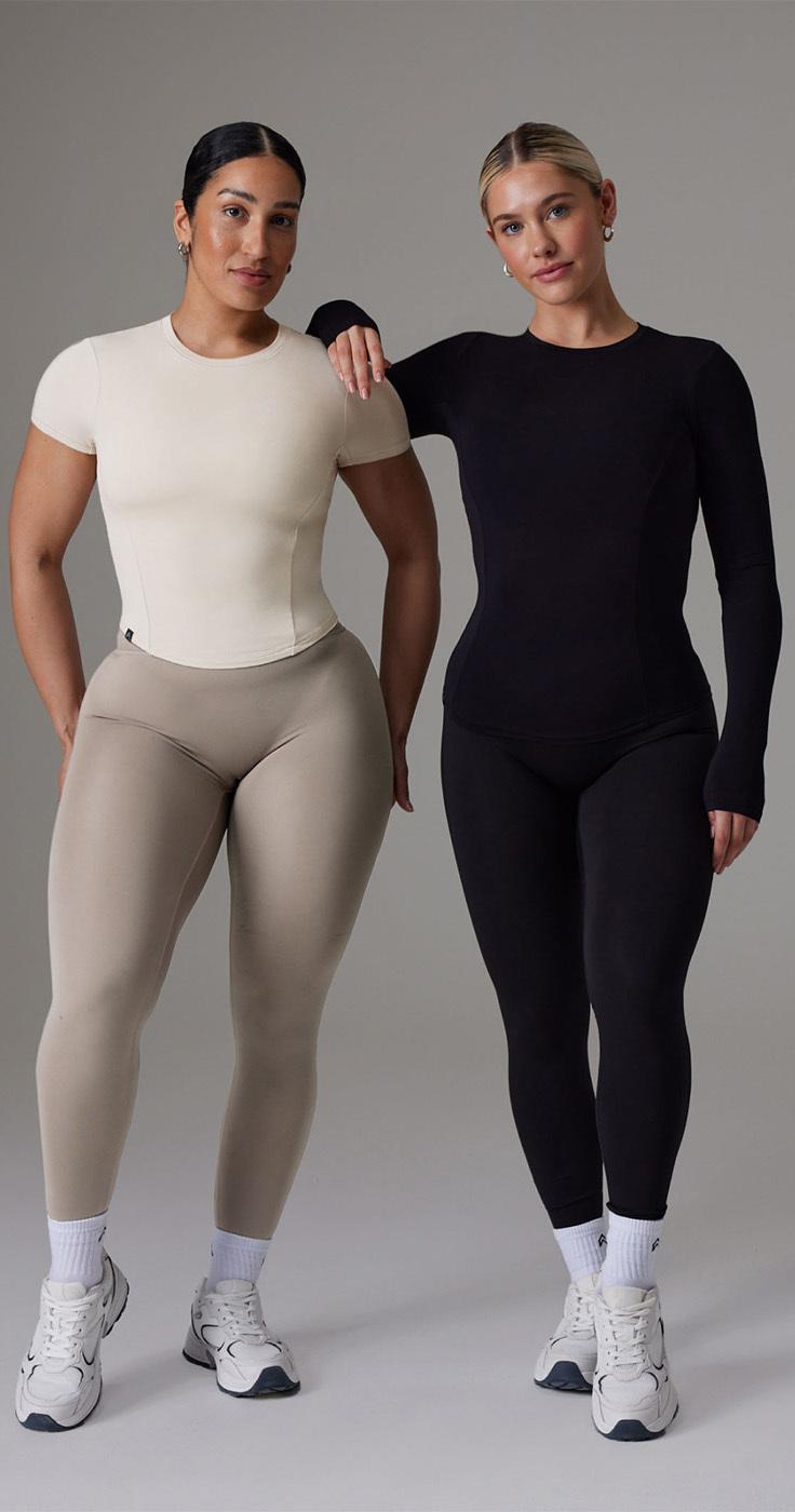 Two women standing side by side, both wearing Oner Active form-fitting workout clothes. The woman on the left is in a beige top and light brown leggings, while the woman on the right is in a black long-sleeve top and black leggings. They stand against a plain grey background.