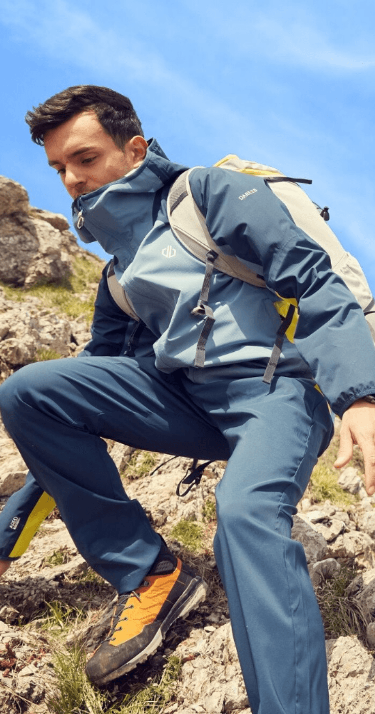 A person wearing a blue hiking outfit and backpack climbs a rocky hill under a bright blue sky. In the background, another individual stands atop the rocks, enjoying the scenic view.