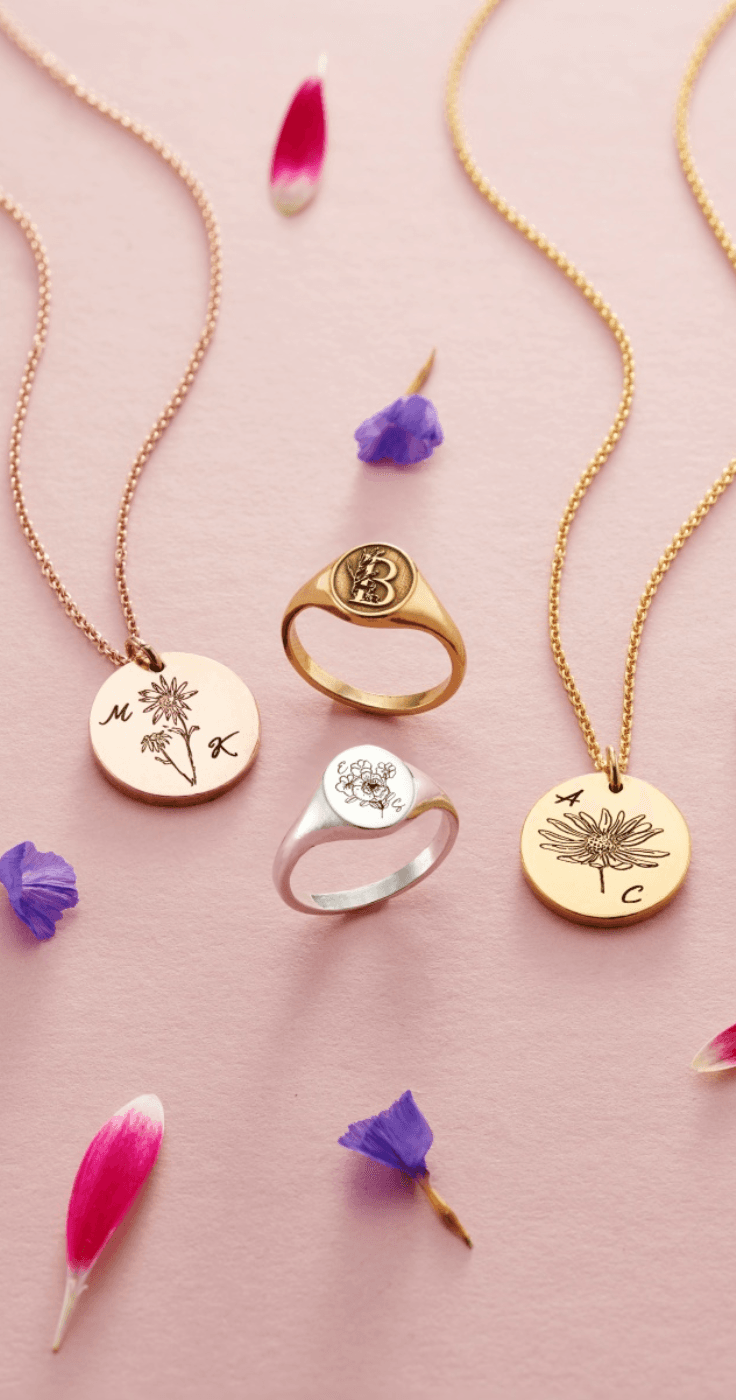 Personalised jewellery from Posh Totty featuring gold and silver signet rings and engraved floral initial necklaces, displayed with delicate petals on a pink background. Engraved Jewellery Gifts for Disabled Women.