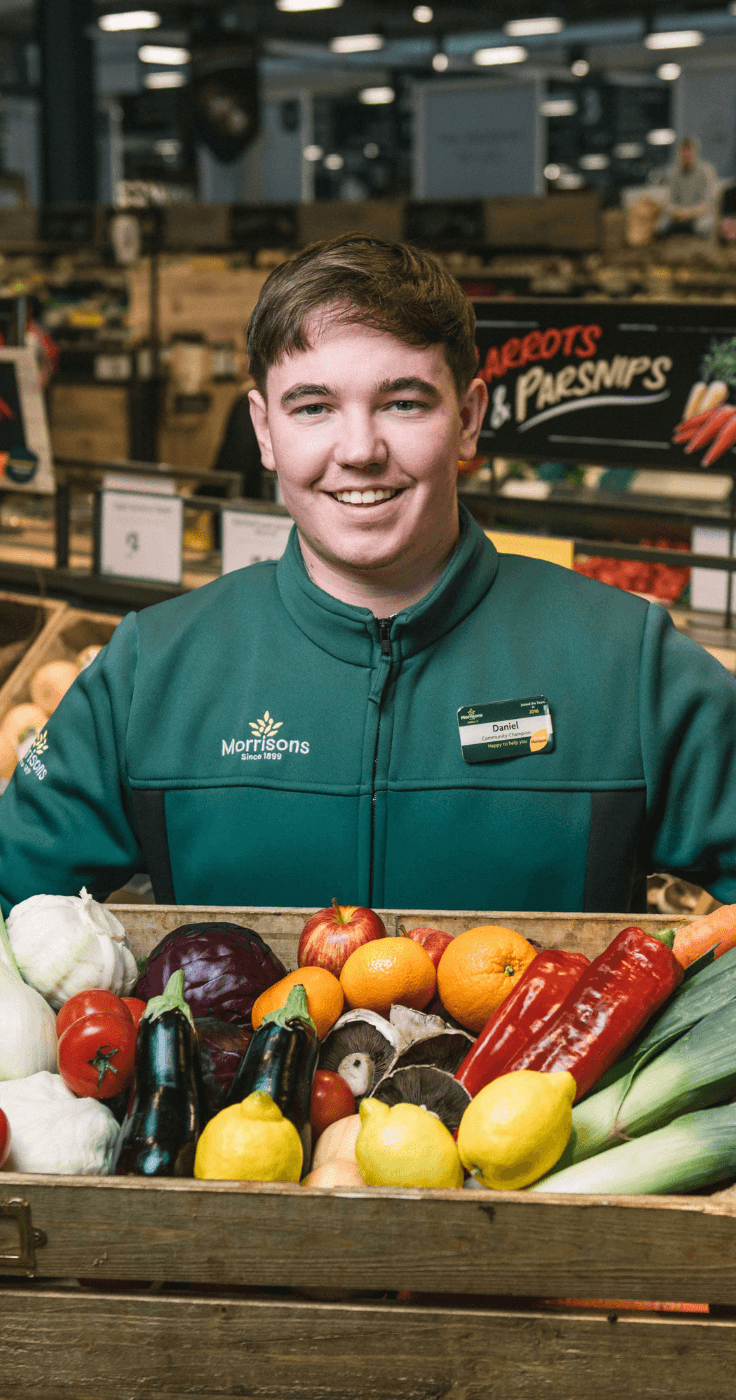 A smiling Morrisons staff member holding a crate of fresh vegetables and fruits in the produce section. Showcasing Purpl's disabled discounts for affordable, high-quality groceries, making everyday shopping accessible in the UK.