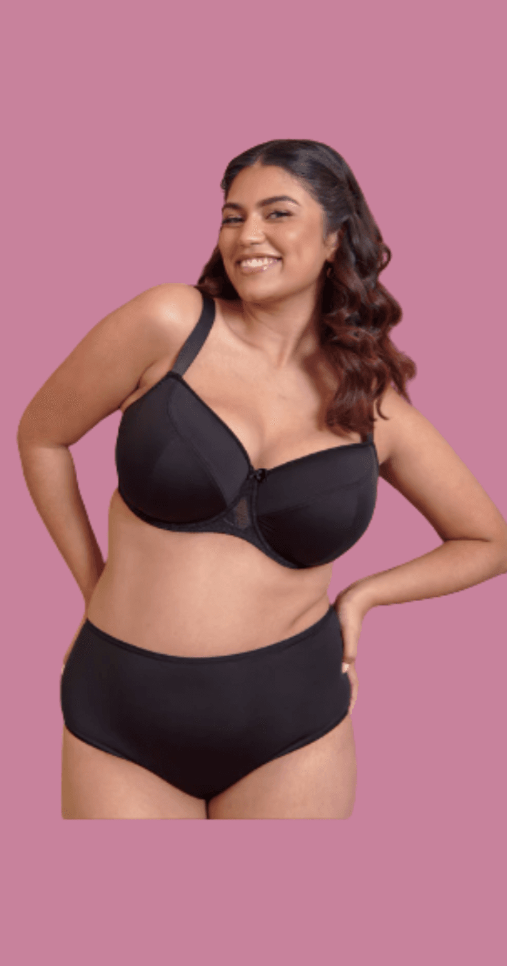 Woman smiling and posing in a black supportive bra and matching high-waisted briefs against a pink background. Plus size lingerie discounts, affordable supportive bras for disabled women, inclusive underwear offers UK.