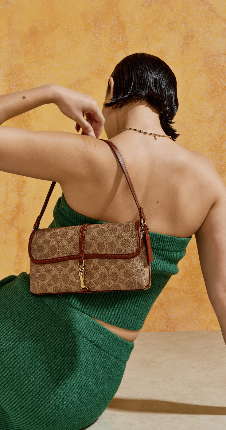 Stylish woman in a green knitted outfit carrying a Coach shoulder bag with classic monogram print, perfect for fashion lovers looking for designer handbag discounts and luxury accessories offers.