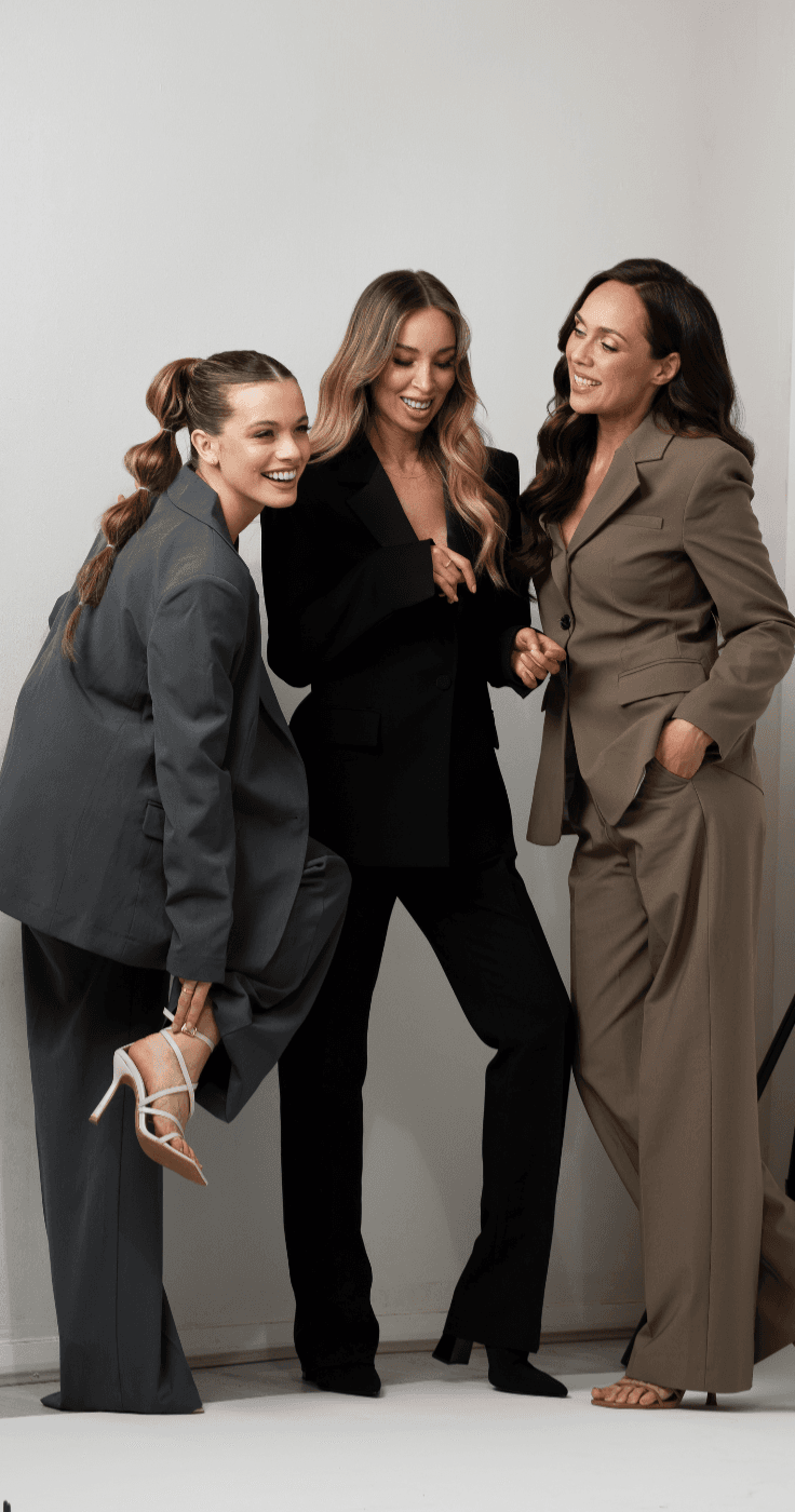 Three women dressed in smart suits laughing together in a studio, one lifting her foot to adjust her heel, promoting confidence and connection through Hair Rehab London with inclusive beauty offers for disabled people across the UK.