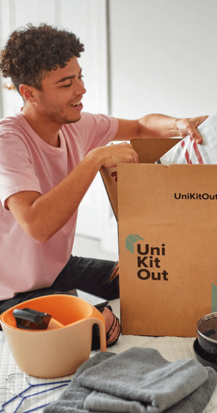 Close-up of a man in a pink T-shirt unpacking a Uni Kit Out branded box, pulling out a kitchen item, with other moving boxes in the background, showcasing affordable home starter packs UK, kitchen starter set discounts, and disabled student moving offers.