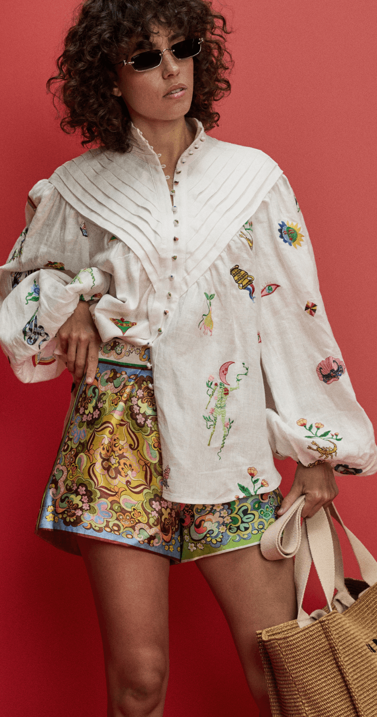 Stylish woman wearing a white embroidered blouse with statement sleeves and colourful patterned shorts, paired with sunglasses and a woven handbag, showcasing luxury designer fashion available at Coggles with exclusive clothing discounts and premium style offers.