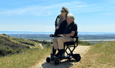  A woman stands next to an older man seated on a mobility scooter on a dirt path atop a hill. Both are dressed in casual clothes and sunglasses. The horizon shows a clear sky and a blur of distant landscape beneath them. They appear to be enjoying the scenery.