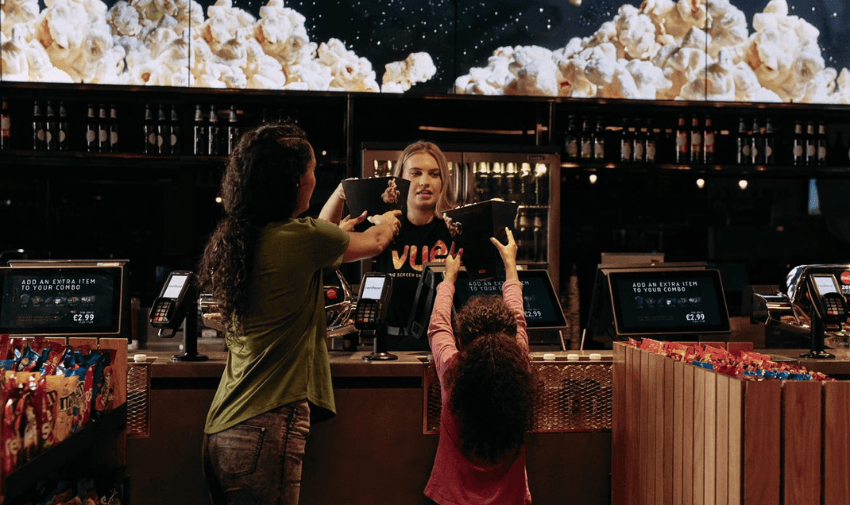 A child reaching for popcorn at the Vue cinema counter while her parent is served by a friendly staff member, highlighting inclusive family cinema experiences and affordable Vue Offers for Disabled People.