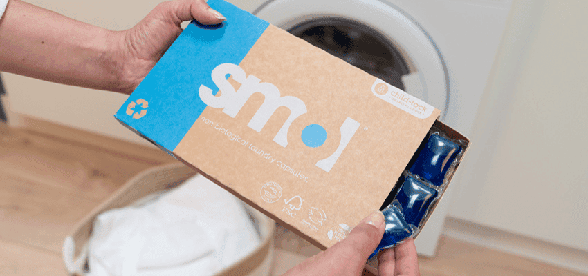 Hands holding smol laundry capsules box next to washing machine promoting affordable household essentials with disabled discounts in the UK