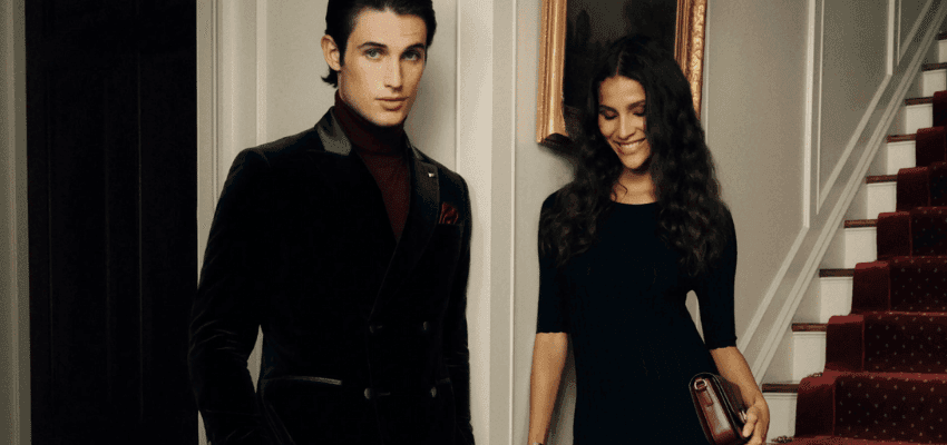 A man in a dark velvet suit stands beside a woman in a black dress inside a stylish hallway with a staircase behind them. Premium fashion image promoting Purpl discounts for people with disabilities and long term health conditions, supporting accessible clothing options.