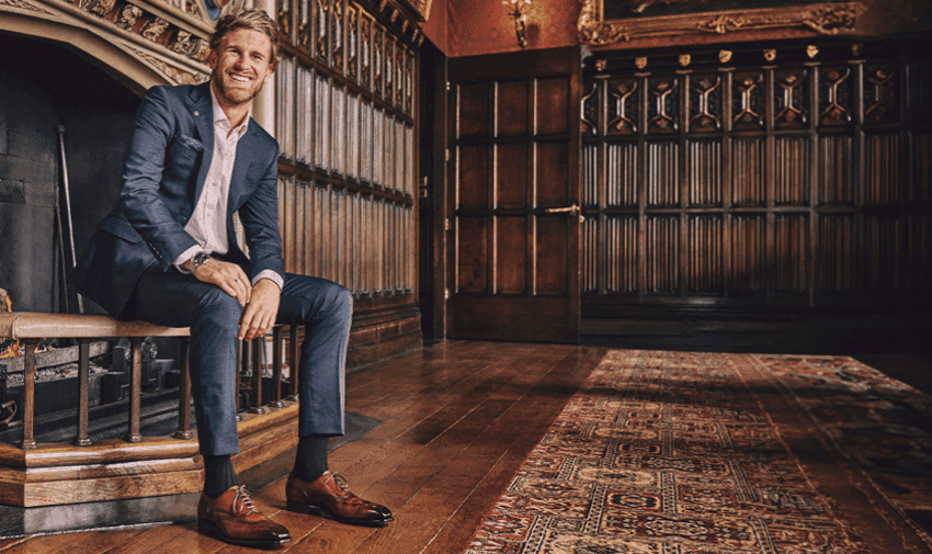 A man in a suit and smart brown shoes sits on a bench near a fireplace in a room with dark wooden paneling and an ornate rug
