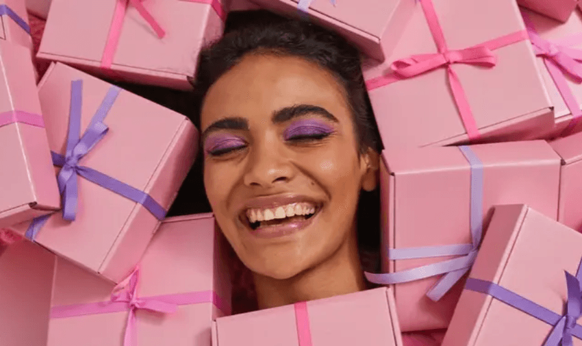 Woman smiling with eyes closed while surrounded by pink ribbon-wrapped gift boxes. Not On The High Street offers feel-good gifting with disabled discounts and personalised deals.