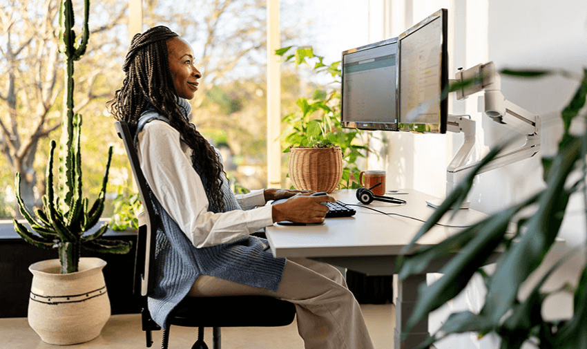 Black woman with long braids working from home at a white ergonomic desk and dual-screen monitor setup, sitting comfortably in a modern chair with natural light and indoor plants, highlighting inclusive workspace products and practical home office discounts for disabled people in the UK.