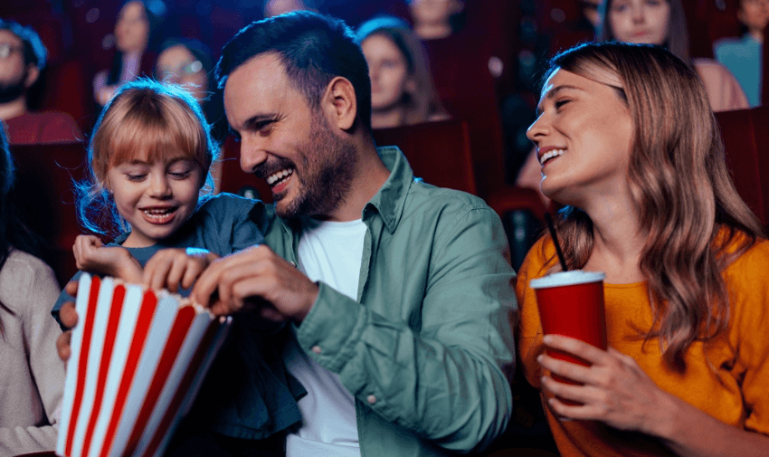 Happy family enjoying popcorn and drinks at the cinema, with a child reaching into the bucket. Perfect for promoting cinema savings for families and mobility discounts and offers for carers in the UK.