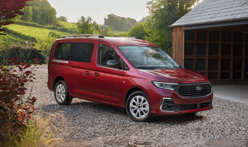 A red Ford MPV parked on a gravel driveway beside a wooden barn, surrounded by greenery, representing spacious and practical vehicles ideal for mobility conversions or family use with accessibility features.