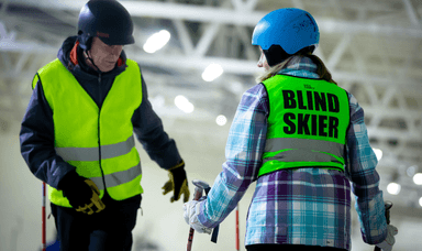 A person wearing a blue helmet and a vest labeled "Blind Skier" holds ski poles while talking to another person in a black helmet and yellow vest. They are standing indoors on a snowy surface.