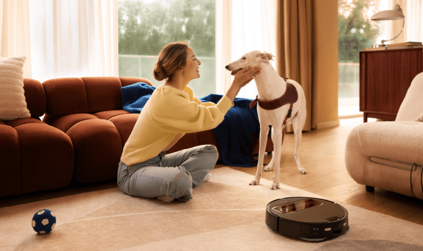 A woman smiles at her white dog while a Roborock vacuum works quietly on the carpet, showing the ease of pet care with smart cleaning. Ideal for accessible home tech discounts for disabled people living independently with pets.