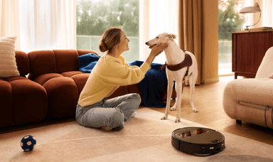 A woman smiles at her white dog while a Roborock vacuum works quietly on the carpet, showing the ease of pet care with smart cleaning. Ideal for accessible home tech discounts for disabled people living independently with pets.