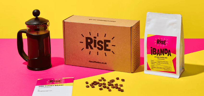 Brightly styled RISE Coffee Box setup with a cafetière, coffee beans, and a RISE Ibanda coffee bag against a pink and yellow background. Promotes sustainable coffee gifts and subscription offers for Purpl members, including exclusive disabled member discounts on eco-friendly, small-batch coffee deliveries.