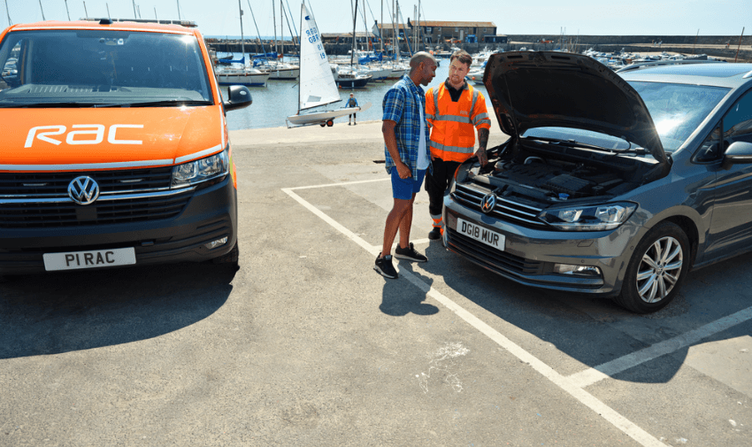 An RAC roadside engineer assists a driver with a broken-down car near a harbour, with both standing beside an RAC van and open car bonnet, representing accessible breakdown cover and inclusive support for disabled motorists across the UK