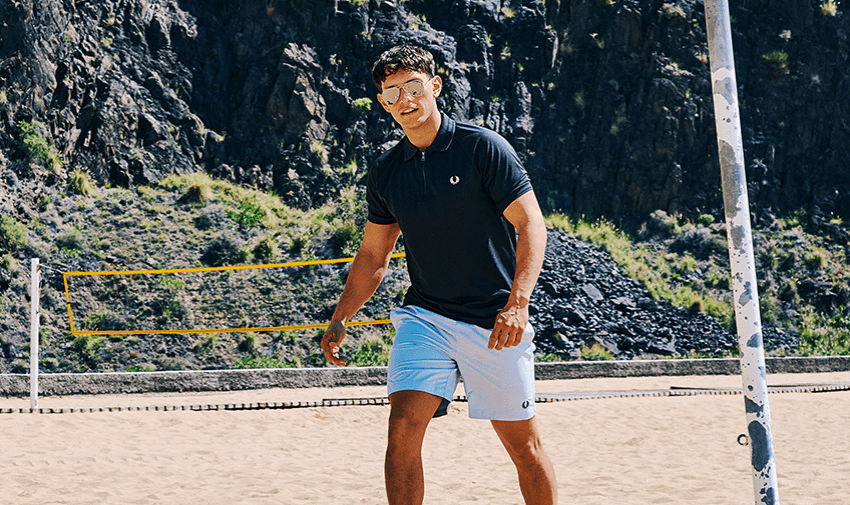 Young man wearing a navy Fred Perry polo and light blue shorts playing volleyball on the beach. Disabled fashion discounts and adaptive menswear offers for active lifestyles.