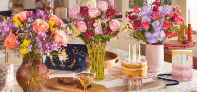 Colourful birthday flower arrangements with roses, gladiolus, and mixed blooms in glass vases, displayed beside a birthday cake and card on a bright table. Keywords: Bloom and Wild Birthday Flowers, Send Flowers Online UK, Birthday Bouquets with Disabled Discounts, Celebration Flowers UK.