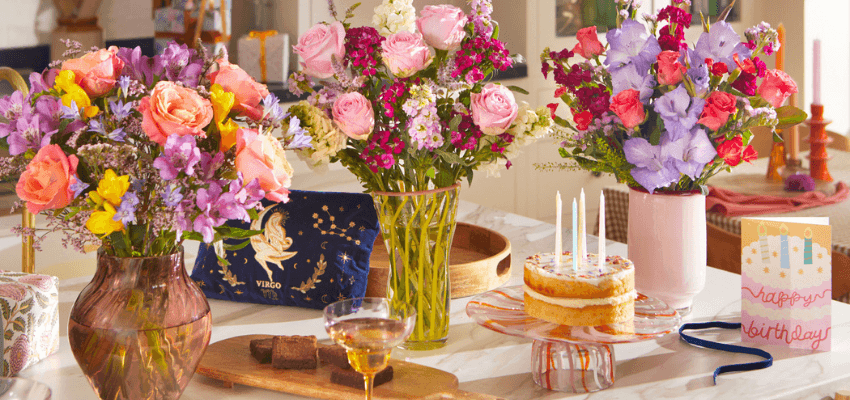 Colourful birthday flower arrangements with roses, gladiolus, and mixed blooms in glass vases, displayed beside a birthday cake and card on a bright table. Keywords: Bloom and Wild Birthday Flowers, Send Flowers Online UK, Birthday Bouquets with Disabled Discounts, Celebration Flowers UK.