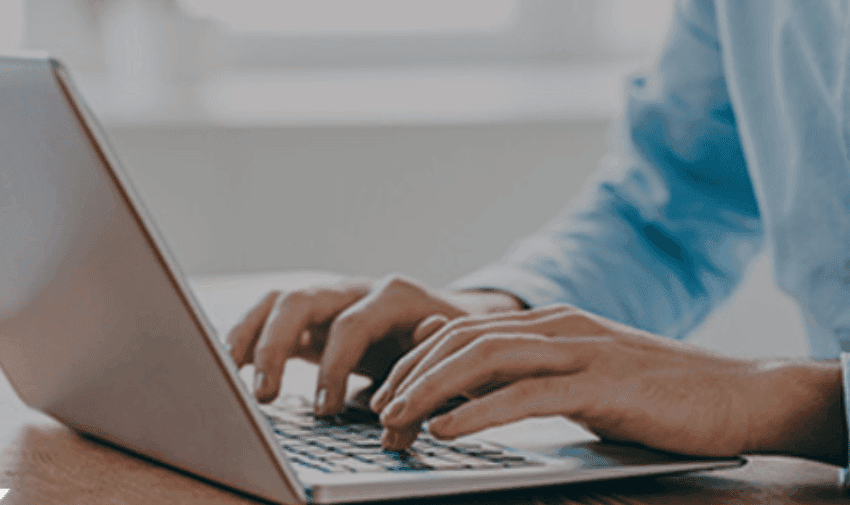 a person typing on a silver laptop whilst sat a wooden table