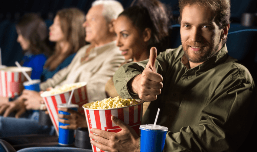 A smiling man giving a thumbs up while seated at the cinema, surrounded by friends and family with snacks. Great for promoting accessible movie discounts and group-friendly cinema deals for disabled people.
