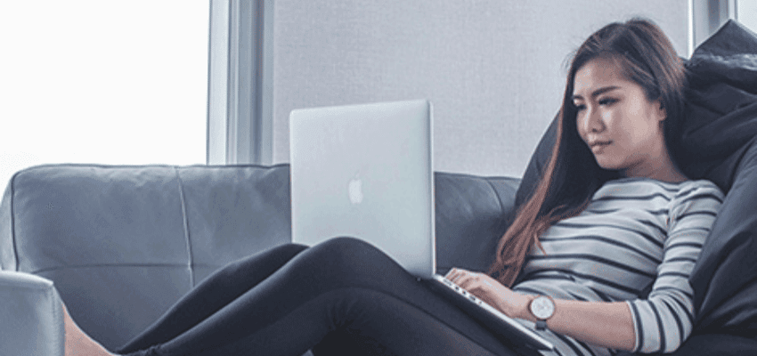 A woman with long hair sits on a grey couch, wearing a striped long-sleeve shirt and black leggings. She has a neutral expression and is focused on her open laptop, which is resting on her lap. She appears to be in a modern, well-lit room with large windows.