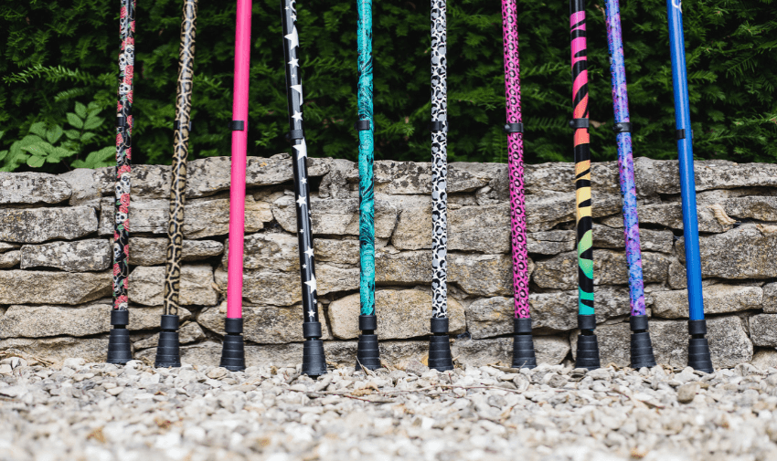 A vibrant line-up of Cool Crutches and walking sticks with bold animal prints, bright colours, and graphic patterns, displayed upright in front of a rustic stone wall — representing stylish and accessible mobility aids.