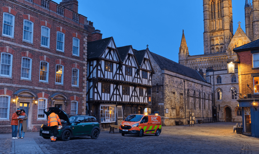 A roadside assistance worker helping a couple with their broken down car in a historic town square at dusk