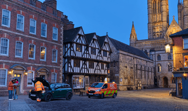 A roadside assistance worker helping a couple with their broken down car in a historic town square at dusk