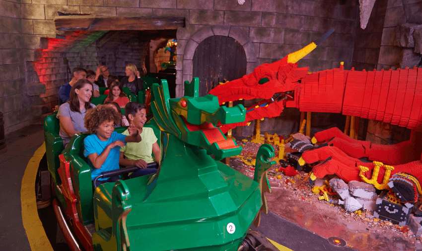 Visitors on a dragon-themed ride inside Legoland, passing a large red LEGO dragon in a medieval-style setting with stone walls. Shopping Discounts for Disabled People and Family Attractions Deals UK.