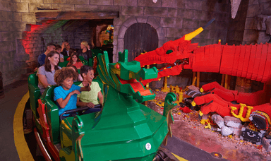 Visitors on a dragon-themed ride inside Legoland, passing a large red LEGO dragon in a medieval-style setting with stone walls. Shopping Discounts for Disabled People and Family Attractions Deals UK.