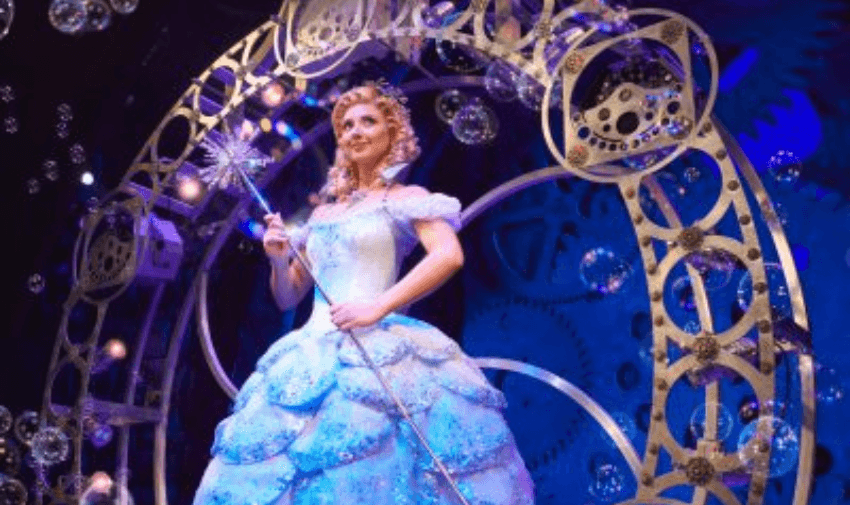 Performer dressed as Glinda the Good Witch in a sparkling blue ball gown, holding a wand and surrounded by bubbles on the set of Wicked, showcasing accessible West End theatre experiences and disabled entertainment discounts in the UK.