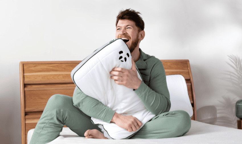 Man in green loungewear sitting on a bed, smiling and hugging a white panda-branded pillow, promoting sustainable and comfortable bedding.